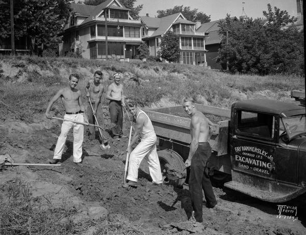 Five University of Wisconsin-Madison athletes (L. to R. Walter Gnabah, Russel Rebholz, George Wright, McClure Thompson, and William Eller) shoveling dirt at Camp Randall with a Fay Hammersley, Jr. Excavating truck and houses on Breese Terrace in the background.
