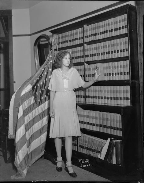 Queen contestant, Disabled American Veterans Ball, standing by bookshelves in front of flag.