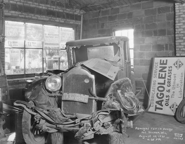Damaged Packard sedan in an auto garage with Tagolene oils and greases sign.