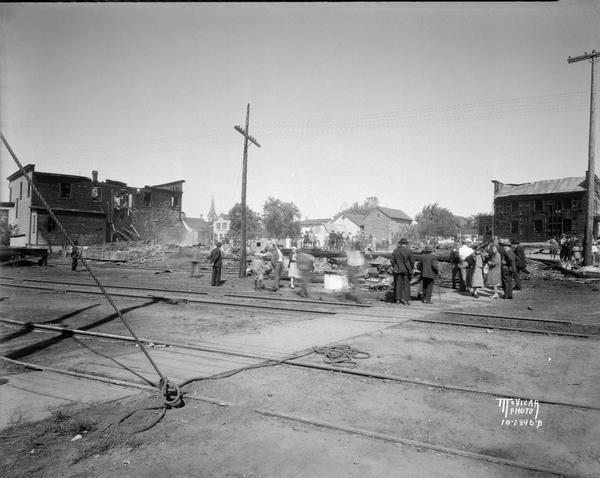 Ruins from fire. There are railroad tracks in the foreground, and people standing and looking at the damage.