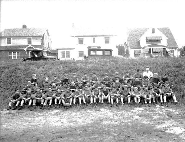 Outdoor group portrait of the West High School football team. Houses are on a hill in the background, and automobiles are parked in front of them.