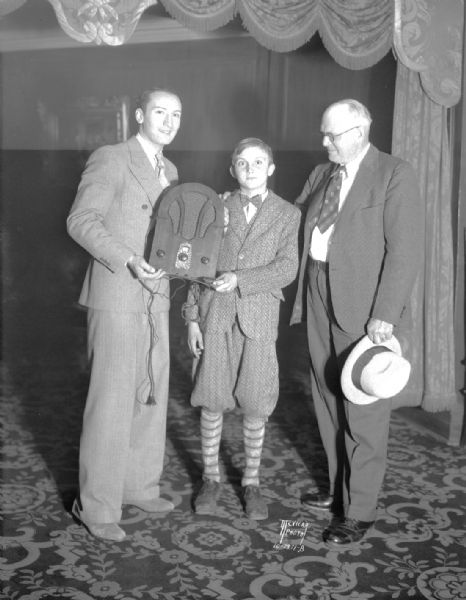 Two men are presenting a radio to a boy, Phillip Harris, as an award for naming the Vilas Zoo lion cubs. The names were "Thor" and "Yama." Left to right in the picture are: Irving Edwards, Master of Ceremonies of the vaudeville show at the RKO Orpheum theater, Phillip Harris, Fred Winkelemann, Director of the Henry Vilas Zoo (Vilas Park Zoo).