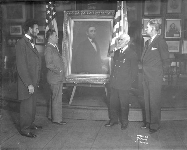 Abraham Lincoln impersonator, Charles E. Bull, Hollywood, California and Jesse S. Meyers, commander of the Madison G.A.R. post, and two other men are standing in front of a portrait of Lincoln in the G.A.R. room in the Wisconsin State Capitol.