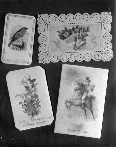 Four antique Christmas cards. At the top left is a copper engraved robin card, 1840. Top right is a harp and rose greeting card hand-painted in 1800. Lower left is a homemade card of pressed ferns and flowers, 1885. Lower right is a rare example of first printed card showing rider carrying corsage, 1850.