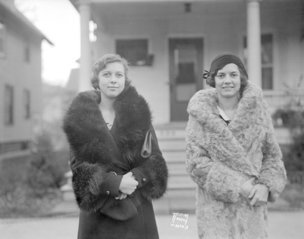 Gretchen Smoot, U.W. Co-ed and cousin of Utah Senator Reed Smoot, and another woman, both wearing fur coats, standing in front of a house.