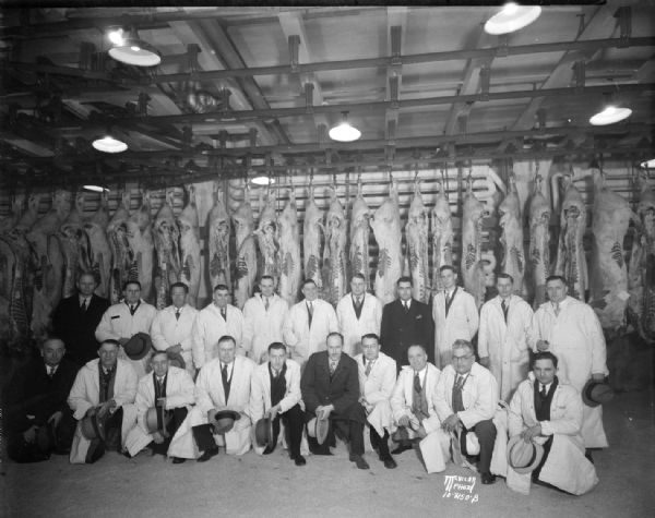 Group portrait of Oscar Mayer salesmen taken in front of sides of beef hanging in the cooler.