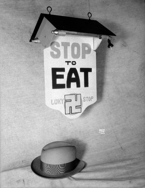 "Stop to Eat, Luky (and a swastika symbol) Stop" sign, with a man's fedora hat resting below. Taken for Mr. Green.