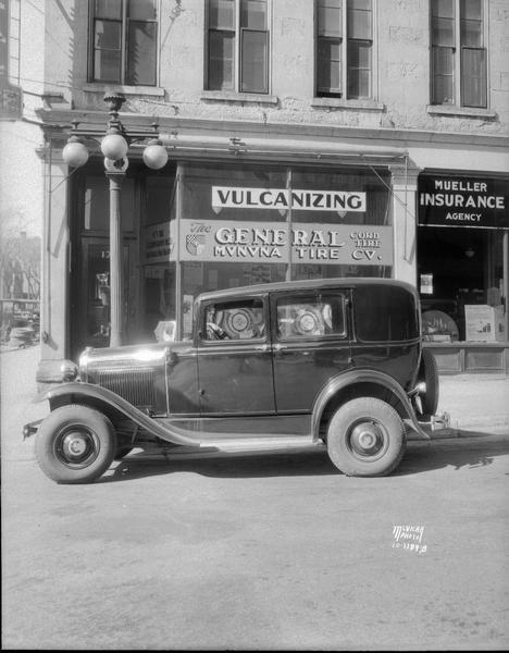 General "Doughnut" tires on Ford in front of Monona Tire Co., 128 S. Pinckney Street. There is a sign for the Mueller Insurance Agency on the left at 126 S. Pinckney Street.
