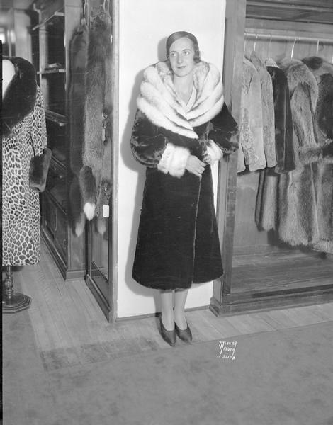 Woman modeling a fur coat at Kessenich's store in the fur coat department. Other coats are displayed on racks and stands on the left and right.