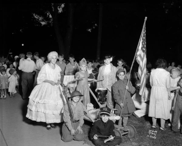 Group portrait of children dressed as George and Martha Washington, and Liberty riding in a chariot and soldiers, one of which is carrying a flag for a Lantern Parade.