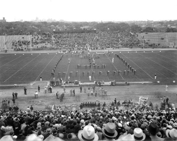 Elevated view from stands of the University of Wisconsin-Madison Marching Band in "I" formation on the Camp Randall football field for U.W. - Iowa game. The Wisconsin State Capitol is in the distance.