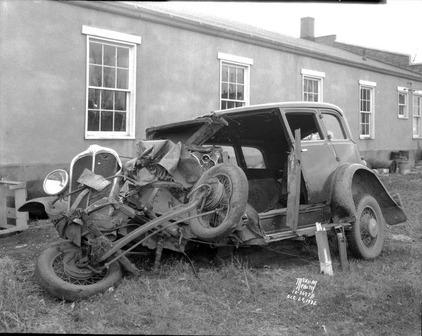 Wrecked Oakland Sedan, from the front left. There is a building in the background.