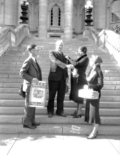 Chief of Police William McCormick buying the first Forget-me-not from the Disabled American Veterans on the Wisconsin State Capitol steps. Left to right: L.J. Kjer, state chairman holding sign, Chief McCormick; Viola Hansen, sales chairman, pinning on the flower, and Evelyn Anderson.