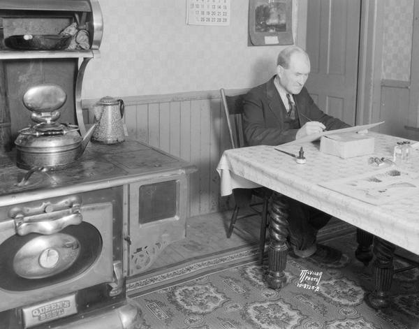 Tom Brown, cartoonist, drawing at his kitchen table beside a large Queen's Best wood burning stove.