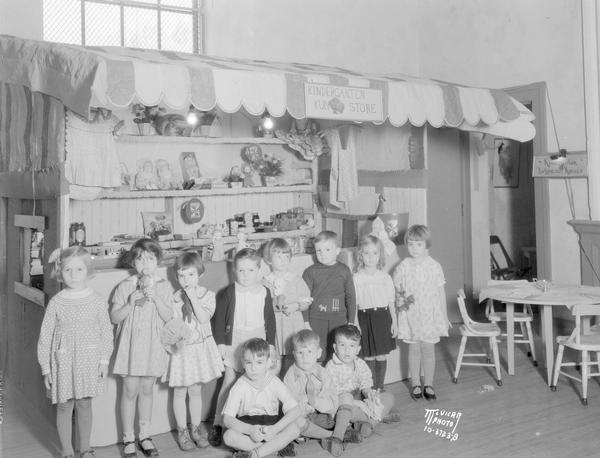 Edgewood kindergarten country store setup. Sign says: "Kindergarten Kun(tree) Store" (rebus), with group portrait of eleven children.