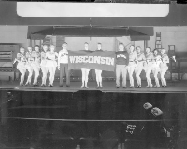 Fanchon & Marco girls, graduates of Carla Tormey School of Dance, Milwaukee, rehearsing a Homecoming show with University of Wisconsin cheerleaders, Joe Stascoo and Bill Stuewe, on the Orpheum stage. Fanchon & Marco were a brother and sister choreography duo who toured the country with variety shows in the 1920s and 1930s.