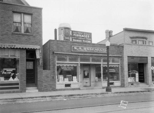Exterior of Edward Brandes Heating Co., with a Premier DeLuxe furnace perched on the roof of store, and Sven A. Rosendahl, plumber, 2211 Atwood Avenue. Also shows Carl M. Johnson, tailor, at 2209 Atwood Avenue, and Seeliger & Stumpf Grocery and Meats (Hormel Plan Market) building at 2213 Atwood Avenue.