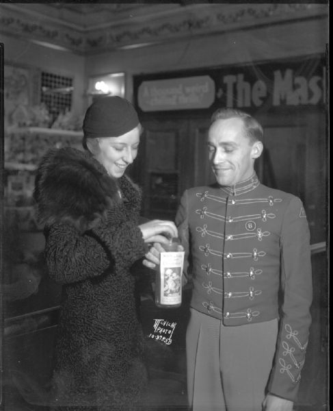 Dorothy Hoffman, member of Phi Sigma Sigma sorority, is shown dropping money into milk bottle held by Harley Droster, an usher at the Orpheum Theatre. Money is being collected for the Wisconsin State Journal's Empty Stocking Club. The bottle has a label showing a picture of children and the phrase: "Will you help us to enjoy Christmas?"