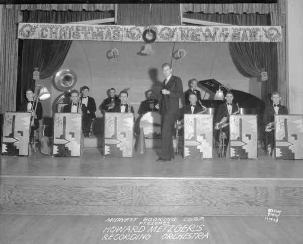 Howard Metzger leading the Howard Metzger's recording orchestra who are sitting on a stage behind music stands, holding instruments. The stage is decorated with a "Merry Christmas, Happy New Year" banner. Printed on negative: "Midwest Booking Corp. presents Howard Metzger's Recording Orchestra."