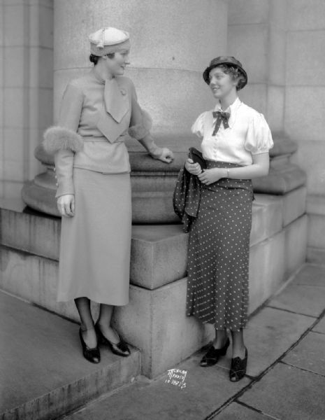 Jean Glanville and Nancy Hotchkiss posing at the Wisconsin State Capitol entrance, modeling clothing from the Cinderella Shop on the Capitol Square for the "Daily Cardinal."