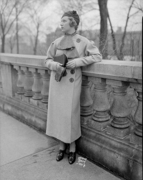 Lois Montgomery, standing next to the railing around the Wisconsin State Capitol, modeling a coat and Fez hat from Baron Brothers Department Store, 14 W. Mifflin Street for the "Daily Cardinal."