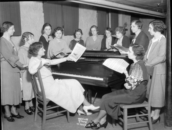 Mrs. Elizabeth Buehler sitting at a piano surrounded by eleven women teachers of the Extension Division of the Wisconsin School of Music, 202 W. Gilman Street.