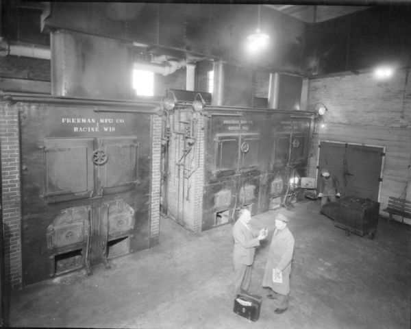 Elevated view of the furnaces in the boiler room at the Dane County Asylum. Two men are standing in the middle of the room.