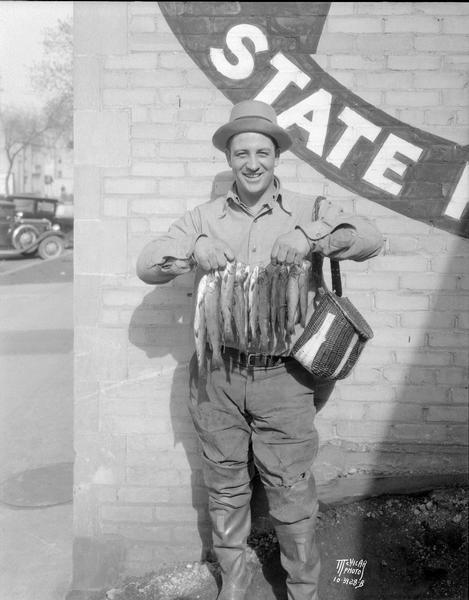 Roy "Chubby" Goodland with a string of trout, caught near Ridgeway. He is wearing waders, has a creel hanging over his shoulder, and is standing in front of a brick wall, which has a sign painted on it that reads: "State."
