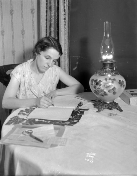 Mrs. Glenn Osmundsen, whose husband works 40 acres of Sunny View farm owned by Howard King, 5710 Lacy Road, sitting at a table with a large decorative kerosene lamp doing bookkeeping for the farm.