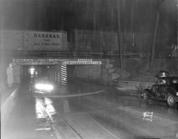 Flooded Park Street viaduct, with a railroad train on tracks above the viaduct. There is a car in the water under the viaduct, and a man is standing on the sidewalk on the left viewing the scene. Sign on viaduct reads: "Danger, Low Clearance 10 Feet 4 in."