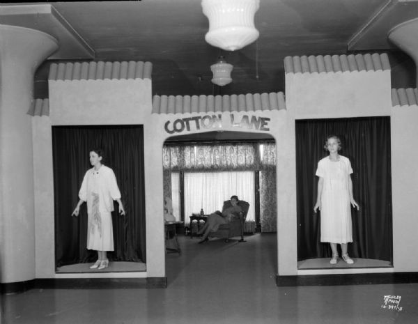 "Cotton Lane" in Hill's Dry Goods Store, 202 State Street, featuring two models, wearing dresses, standing in niches on either side of an arch, with the words "Cotton Lane" framing an open doorway showing a woman sitting in a chair near a window in the background.
