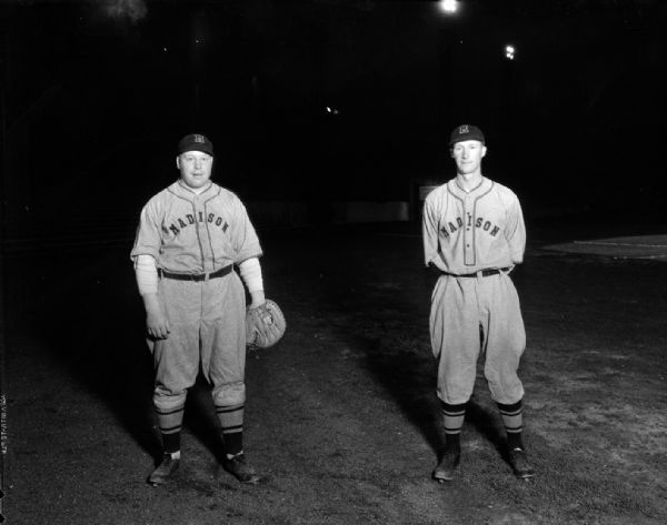 Outdoor portrait of two Madison Blues baseball players in uniform. Richard "Red" Smith with large catcher's glove, and Alvin "Butch" Krueger with his hands behind his back.