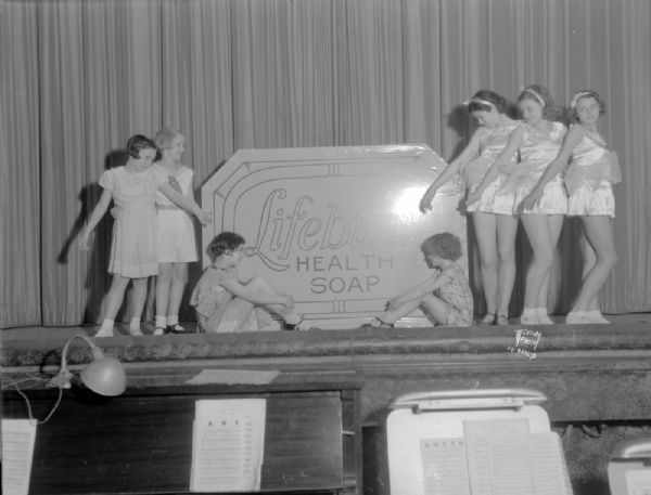 Group of seven dancers posing around an oversize cake of Lifebuoy Health Soap on a stage. The orchestra pit is in the foreground. Billie Usher, Marianne Hebblethwaite, Jacqualine Lorig, Ruth Holt, Jean Deneen, Jane Buellesbach, and Ruthmary Mahoney of the Wheeler Conservatory School of Dancing, perform for the Capital Times Charm School.
