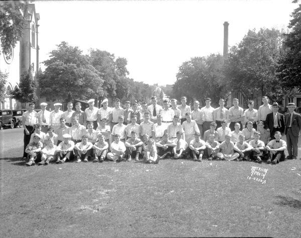 Outdoor group portrait of Capital Times newspaper carriers. In the background is a church building on the left, and a water tower on the right.
