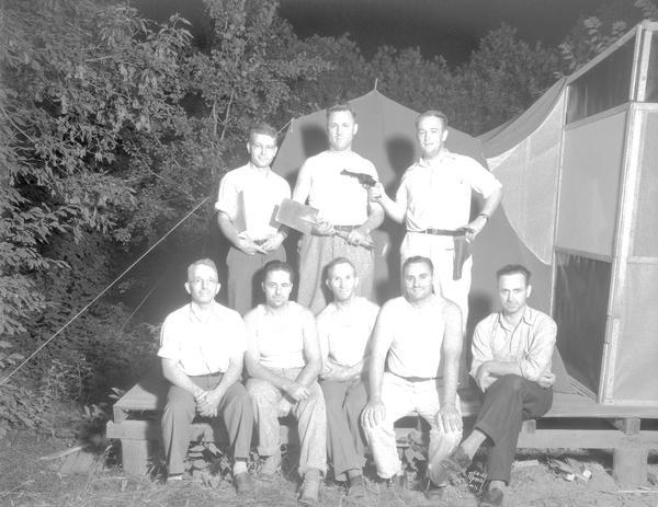 Group portrait of eight male University of Wisconsin tent colony officers. One man is holding a gun, and another is holding a shovel. The camp is also known as Camp Gallistella on the south shore of Lake Mendota, west of second point.