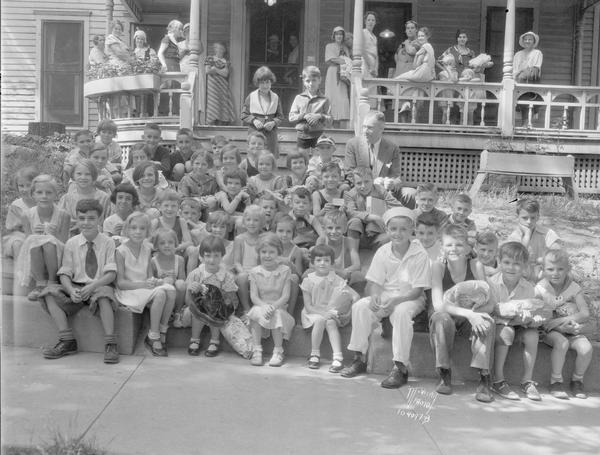 Group portrait of children sitting on porch steps waiting to go to The Capital Times Kiddie Camp. One man (Evjue?) is sitting with them, and several women are standing on the porch.