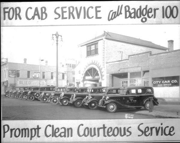 Taxicabs lined up in front of City Car Co., 214 E. Washington Avenue, corner of N. Webster Street, with advertising banners at the top that read: "For cab service call Badger 100," and at bottom: "Prompt Clean Curteous Service."