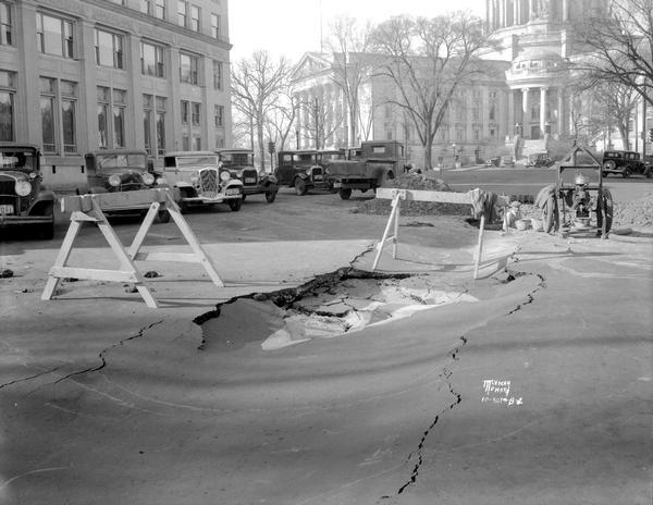 Cave-in on Monona Avenue, caused by water main leak, looking towards Wisconsin State Capitol. View shows automobiles parked in the street, and the State Bank of Wisconsin on the left.