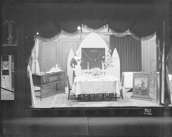 Thanksgiving dining room display window at Burdick & Murray, 15-19 East Main Street. Featuring a mannequin wearing a dress and an apron, linens, china, crystal, and silverware from E.W. Parker Jewelers.