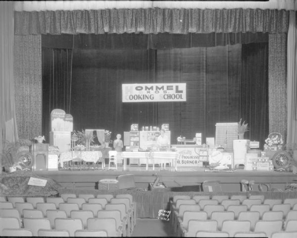 View from center aisle of Hommel's Cooking School set up on the Parkway Theater stage, 6-10 W. Mifflin Street. Displayed is a layout of the cooking demonstration as well as advertising for Johnston biscuits and crackers, Scott towels, Philco radios, Goodyear tires, and Karasnan rugs.