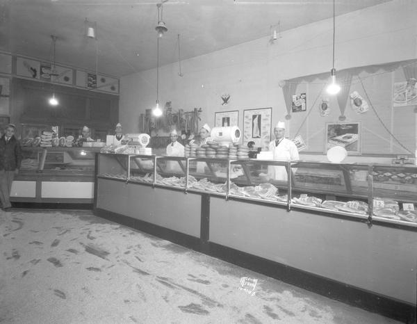 B. Elmer Moore's Cash (meat) Market, 2333 East Washington Avenue, interior. Five clerks are standing behind the counter, and another man is standing in front of the display case on the far left. There is sawdust on the floor.