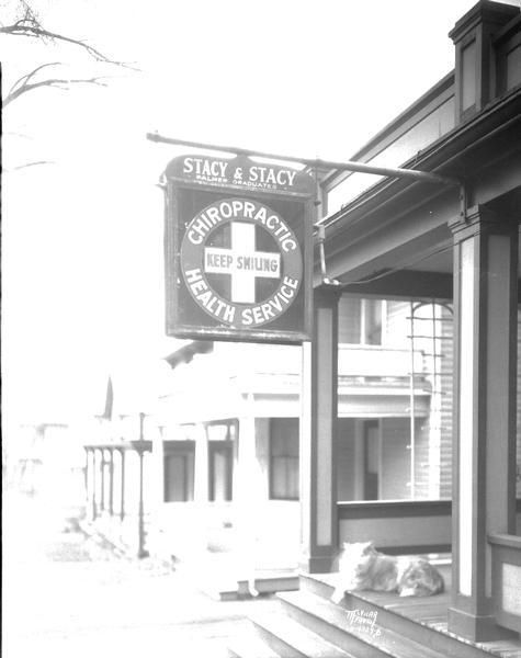 Sign hanging from front porch for Stacy and Stacy Chiropractors, 332 W. Washington Avenue, J. Clifford Stacy and Mary E. Stacy practitioners. The sign reads: "Chiropractic Health Service, Keep Smiling." A dog is sitting on the porch.