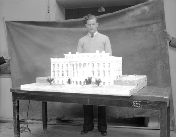 One man is standing behind a cake shaped like the White House, showing the rear view of the White House, baked by the Strand Baking Company, 2007 Atwood Avenue, for the Roosevelt Birthday Ball. Men are holding a backdrop behind the man.