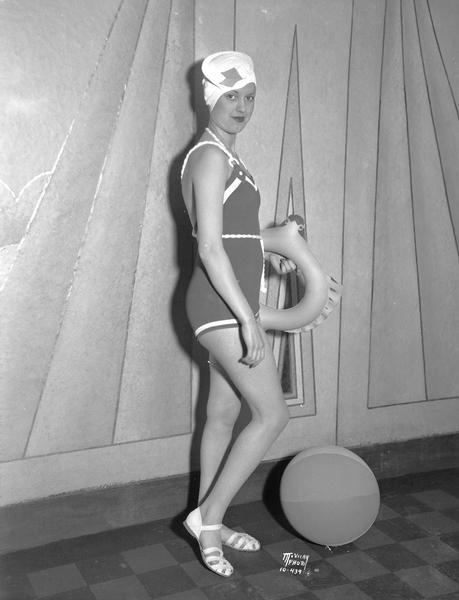 Helen King, side view, modeling a 1934 rubber bathing suit, made by U.S. Rubber Co. She is also wearing a bathing cap and sandals, and is holding an inflatable pool toy. A beach ball is on the floor at her feet.