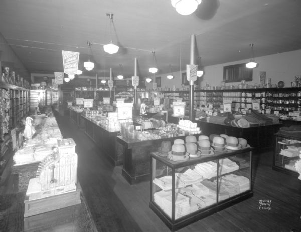 Grand opening of The Fair Store, 1202-1204 Williamson Street. Interior view from the right rear corner. There is a large cash register on the counter in the left foreground. Hats are on top of a display case in the center.