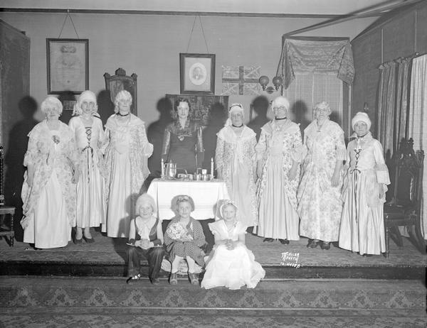 Eight women and three children in colonial costumes on stage at the I.O.O.F. Hall. Rebekah is the women's branch of the Independent Order of Odd Fellows.