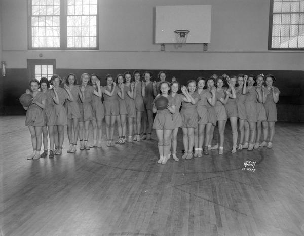 Two lines of girls dressed in gym uniforms waving to the camera, first girl in each line is holding a basketball, in the gym at Nakoma School, 3872 Nakoma Road.