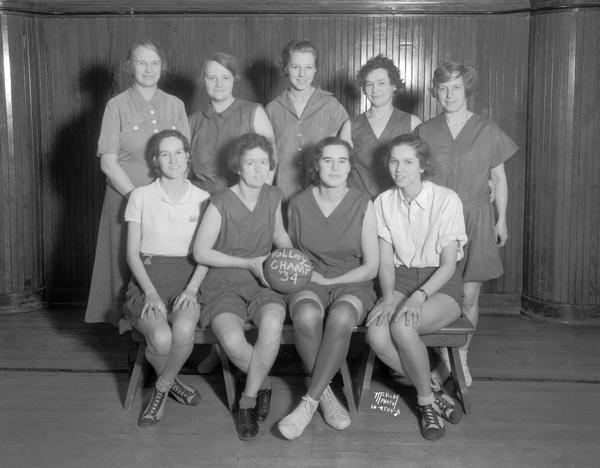 Group portrait of 9 women volleyball players in gym dress holding a ball that says "Volly Champs '34."