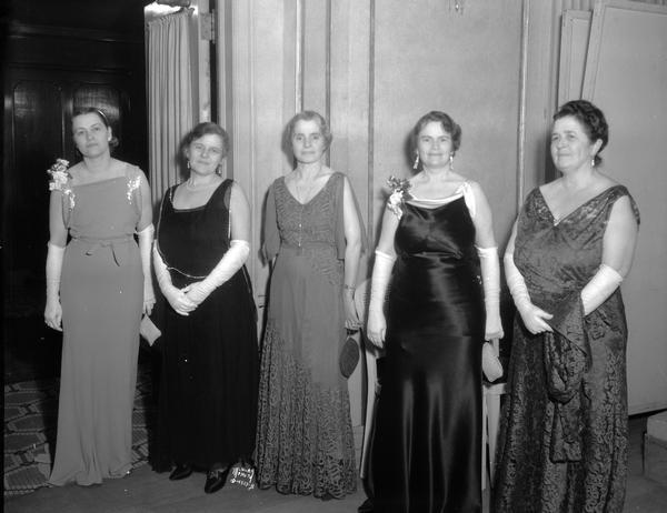 Five women in ball gowns standing in the Attic Angels Ball receiving line in the Loraine Hotel, l to r: Mrs. Erwin Schmidt; Mrs. Wallace W. Chickering, Society President; Mrs. T.M. Priestley; Mrs. Mary Swenson North; Mrs. C.E. Mendenhall.
