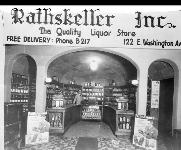 Rathskeller Inc., 122 E. Washington Avenue, liquor store interior, framed with arches, and a clerk behind the counter. Includes an advertising banner that reads: "Rathskeller Inc., The Quality Liquor Store" that has been added to the negative.